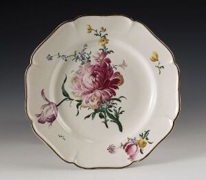  An octagonal-shaped ceramic plate with scalloped edges, featuring a detailed painting of a large pink rose in the center, flanked by smaller flowers and foliage in shades of purple, yellow, pink, and green, against an off-white background. The edge of the plate has subtle brownish-gold accents.