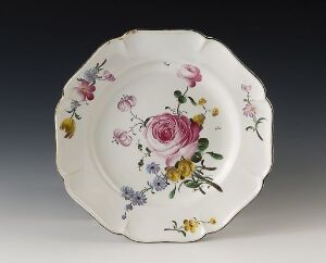  A scallop-edged porcelain plate with a detailed floral pattern featuring a prominent pink rose surrounded by smaller flowers in lavender, yellow, and blue hues against an off-white background.