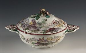  An ornately decorated oval porcelain tureen with a pastoral scene, featuring a vegetable-shaped knob on the lid and burgundy trim, set against a neutral background.