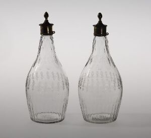  Two clear glass antique bottles with vertical ribbed designs and metal shaker tops on a light grey background.