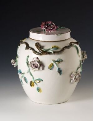  A decorative ceramic jar with intricate three-dimensional floral designs in shades of pink, purple, and green with gold accents, set against a neutral gray background.