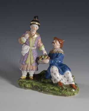  A porcelain figurine entitled "Chinesergruppe," depicting two stylized individuals on a green base, with one standing in a striped purple and yellow coat and black hat, and the other seated in a blue floral dress holding a bowl, reflecting a European interpretation of Oriental design.