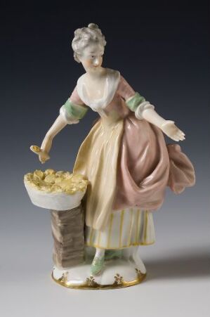  A porcelain figure of a woman in an 18th-century pink, green, and yellow dress, with a draped mauve garment, lifting the skirt to reveal layers underneath, standing next to a pedestal with a bowl of golden yellow flowers or fruit.