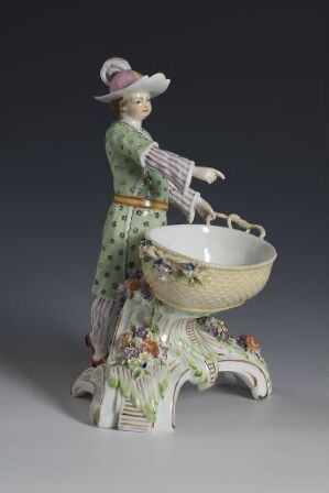  A porcelain figurine depicting a woman in a green, polka-dotted 18th-19th century dress with a lilac hat, mixing in a large bowl, accompanied by a white and gray goat, on a detailed base with floral accents.