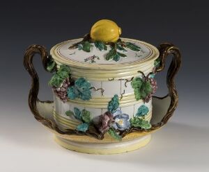  A decorative porcelain soup tureen with a lid, featuring three-dimensional grapevine designs in green and purple, with gold-trimmed handles and a bright yellow lemon as the lid's handle.