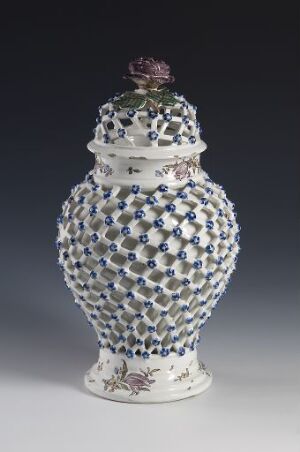  A decorative porcelain pot with a lid showcased against a gradient gray background. The pot has a raised white honeycomb pattern interspersed with blue and green dots and subtle floral designs. The lid is crowned with a purple flower finial.