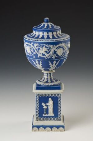  A decorative blue and white porcelain urn with lid on a square pedestal base featuring intricate blue patterns and scenes on a glossy white background, against a neutral gray backdrop. Artistname and title are unknown.