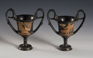  Two ancient Greek kylix drinking cups with black-figure decoration on a neutral grey background. Each cup has a deep bowl, two looped handles, and stands on a thin, circular base. The figures depicted on each kylix are rendered in black with additional red and white details, showcasing traditional Greek pottery techniques. Artistname and title are unknown.