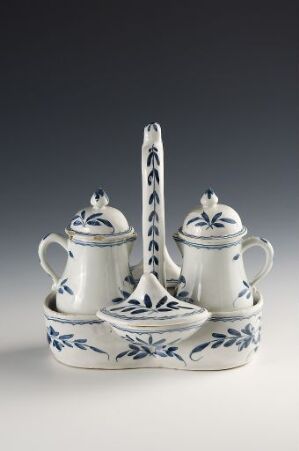  A porcelain cruet stand with a central handle and two covered pots featuring blue floral patterns on a white background, all resting on an ornate, oval-shaped tray with similar detailing, against a soft greyish-blue backdrop.