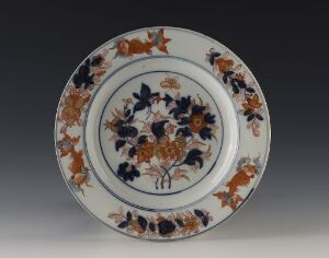  A round, decorative plate featuring a detailed Imari-style design with blue and orange floral motifs and gold accents, set against a neutral gray background.