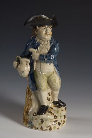  A ceramic mug shaped like an 18th-century man standing on a textured base, wearing a tricorn hat, deep blue coat, cream vest, and beige breeches, holding a grey jug.