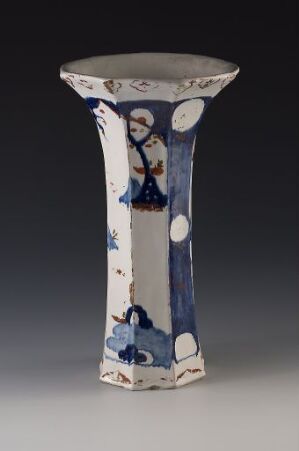  A tall, narrow ceramic vase with a flared top, adorned with cobalt blue and white patterns, featuring floral motifs and an indistinct central scene, against a neutral, dark grey background. Artist and title unknown.