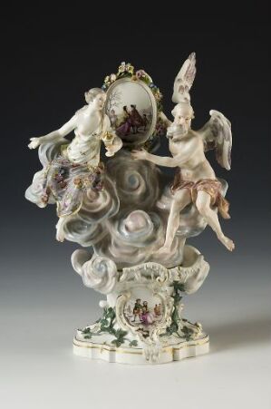  An ornate porcelain figurine on a square base with decorative floral motifs and gilded edges. The sculpture features a cherub with spread wings holding a circular frame around an oval cameo, set against a swirling grayish-white cloud motif.