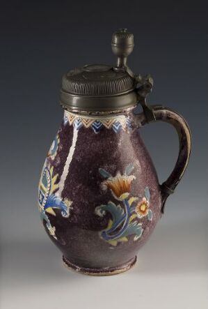  An antique jug with a deep plum body and a decorative floral design in blue, yellow, and orange, featuring a wavy gold band near the neck and a pewter lid on top.