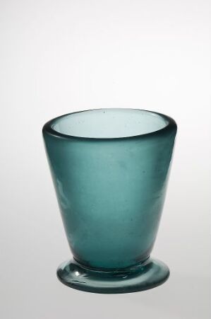  A teal-colored, translucent glass tumbler with a simple design, placed against a light grey background that transitions to a soft gradient near the base.