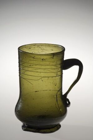  Antique cylindrical glass mug with a green tint and horizontal glass threads, a handle on one side, showing bubbles and imperfections, against a gradient background fading from white to gray.