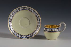  A porcelain cup and saucer set on a gradient gray background. The set is off-white with decorative blue and gold geometric patterns around the rim of the saucer and the edge of the cup, which has a golden interior. The cup has a curved handle with a thin gold outline. Artist name and title are unknown.