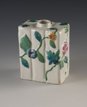  A uniquely shaped ceramic vase with an off-white background color, featuring raised blue, green, and pink floral motifs on its surface, placed against a grey backdrop.