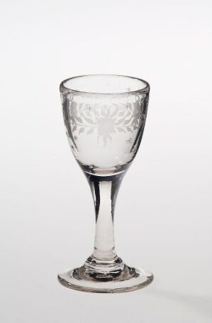  A clear glass goblet with an etched design on the bowl, an elongated stem, and a broad base, set against a light, neutral background.