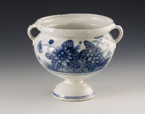  An elegant off-white ceramic bowl with a blue floral pattern and two curved handles, set on a neutral background. The bowl stands on a pedestal foot, and the artwork on the bowl resembles traditional blue and white ceramic design. Artist name and title remain unknown.