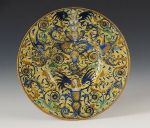  A decorative ceramic plate with a central blue fantastical animal motif surrounded by intricate blue and golden-yellow floral and vegetal patterns on a circular plate.