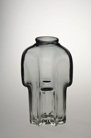  An empty, transparent glass bottle with a design resembling a human figure stands against a neutral background. The bottle's form includes a pronounced neck at the top, broad shoulders, and a base that imitates wide hips, tapering down to a flat bottom for stability.