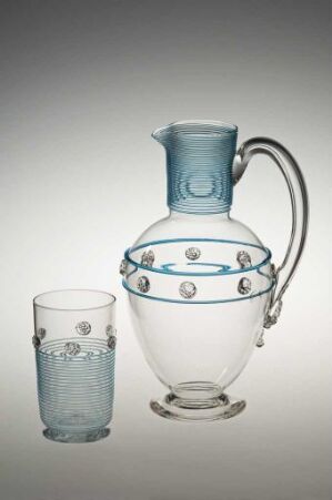  A clear glass pitcher with a frosted band and embossed shield-like designs, along with a matching glass, set against a light grey background that fades into white at the top.