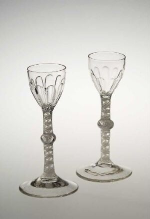  Two transparent glass goblets with decorative heart motifs on the bowls and beaded stems, set against a neutral grey background. Artistname and title are unknown.