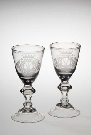  Two clear glass goblets with etched floral designs, side by side against a light grey background, reflecting soft light and casting faint shadows.