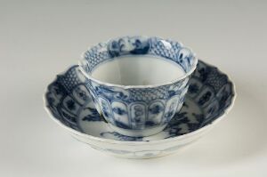  A porcelain tea cup with a blue floral and scroll design sits inside a matching saucer, both on a light grey background, illustrating the intricate cobalt blue patterns typical of traditional East Asian-inspired ceramics.