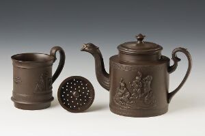  A traditional tea set consisting of a dark brown clay mug, a spherical tea strainer with holes, and a teapot with a serpent-shaped handle and a raised relief design, all set against a neutral background. Artistname and title are unknown.