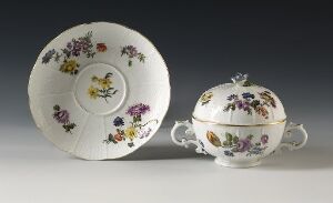  A porcelain tea set with a plate and covered sugar bowl, each adorned with colorful, hand-painted flowers on a white background, set against a neutral gray backdrop.