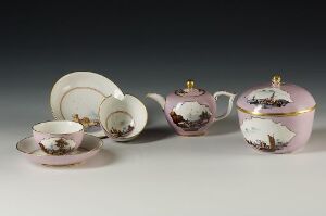  A porcelain tea set with pastoral scenes in pastel colors and gold accents, including a cup with saucer, two teapots, a creamer, and a sugar bowl, arranged against a light gray background.