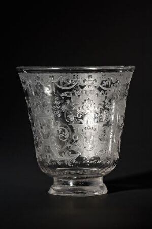  An ornate glass vessel with detailed etchings and scrollwork patterns set against a dark background, highlighting its elaborate design and craftsmanship.