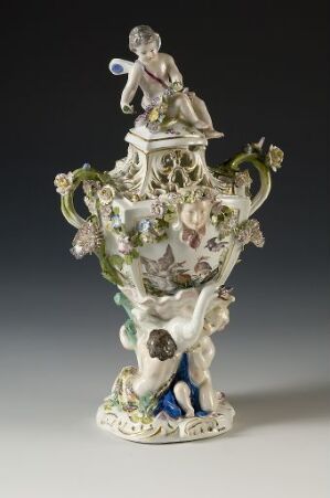  An ornate porcelain ewer with sculped figurines and floral motifs in pastel colors on a dark background. The ewer features a classical female figurine on the lid and is supported by three figures at the base.