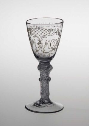  Transparent glass goblet with ornate engravings, including an inscription with the letters "MO", set against a light-to-gray gradient background. The goblet's bowl is decorated with a band of loops, and the stem is fashioned in a twisted column design, leading down to a stable, wide base.