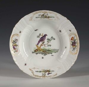  A piece of porcelain tableware from Den kgl. Porcelainsfabrik, featuring a scalloped rim and delicately painted overglaze decoration with a purple and yellow bird perched on a branch in the center, floral motifs, and gold accents, against a creamy white background.