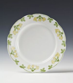  "Maria Nøkleblom" by Porsgrunds Porselænsfabrik AS, a feldspar porcelain plate with a white background and a hand-painted overglaze decoration of green leaves and yellow flowers around the rim, against a soft gray background.