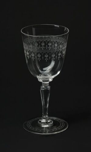  An intricately etched glass goblet with a floral motif stands against a pure black background, visible in shades of gray from the light reflecting off its surface.