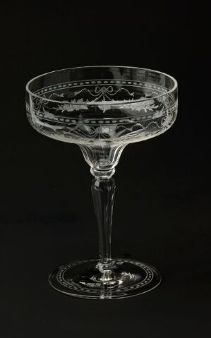  A single clear glass champagne coupe with intricate etching designs on the bowl, standing against a dark black background, highlighting its elegance and craftsmanship.