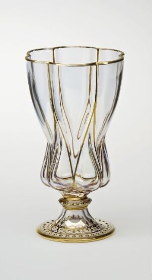  An elegant fluted glass with clear body and gold detailing on the stem and base, featuring vertical ridges and ornate filigree patterns. Artist name and title unknown.