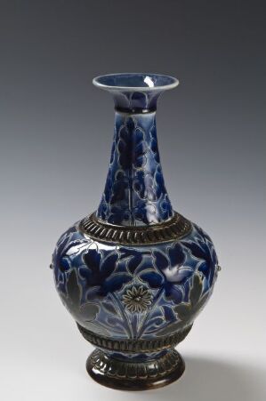  A porcelain vase with a deep indigo blue and white floral design, featuring a slim hourglass shape, metallic trim, and a small emblem, against a gradient gray background. Artist name and title are unknown.