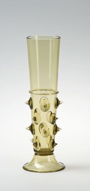  An antique pale yellowish glass vessel with a cylindrical shape, decorated with bulbous protrusions and circular medallions with faces in relief, on a light grey background. Artist name and title are unknown.