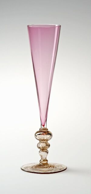  A slender, elegant glass with a soft pink translucent bowl that fades to clear at the rim, atop an ornate stem with gold and clear glass elements, ending in a stable clear base with a gold accent.