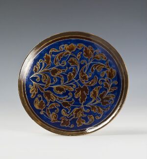  A round decorative plate with a vibrant blue background and symmetrical golden floral designs along the rim, set against a light gray backdrop. Artist and title are unknown.