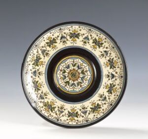  An ornately designed ceramic plate with a pattern of black, gold, and blue floral motifs on a creamy white background, featuring a central medallion with a concentric floral pattern.