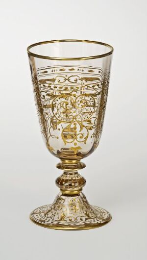  An ornate, baroque style goblet with intricate gold detailing on a transparent glass, featuring elaborate patterns on the bowl, stem, and base, set against a plain background.