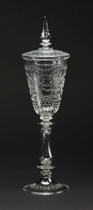  An intricate glass goblet with a pointed lid against a dark background, showcasing detailed patterns etched into its surface and a segmented stem leading to a circular base.