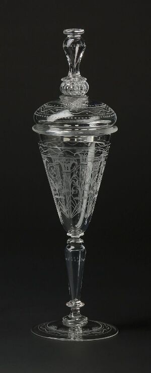  An elegant and intricately designed transparent glass vessel with a cone-shaped bowl and a long stem, topped with a patterned lid and finial, against a stark black background.