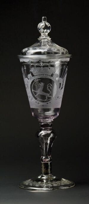  A tall, silver goblet with intricate embossed designs and a figurative finial on the lid, set against a black background. Artistname and title are unknown.